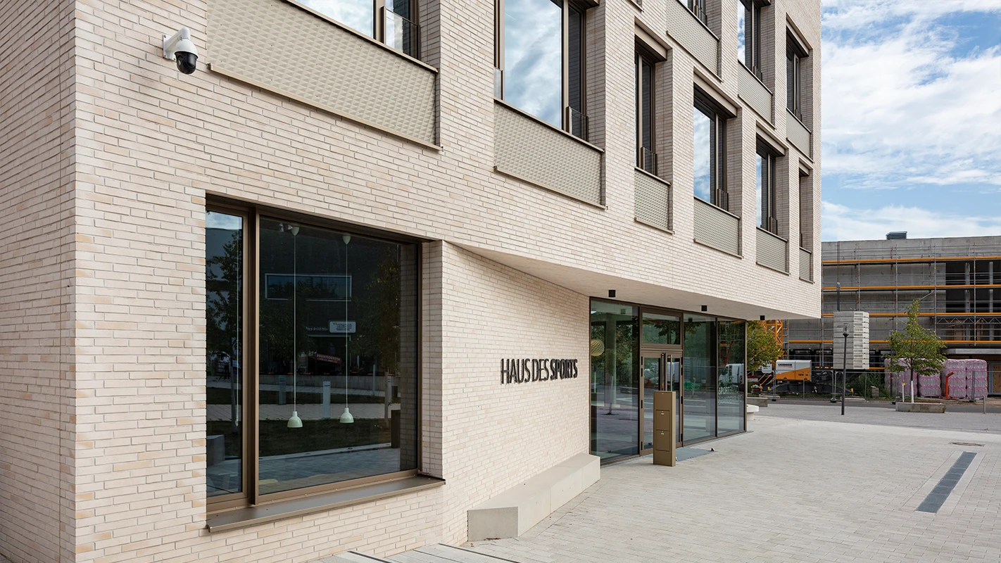 Seitlicher Eingangsbereich des Haus des Sports mit heller Fassadenbekleidung, großen Glasflächen, 3D-Optik-Platten und moderner Fassadengestaltung.