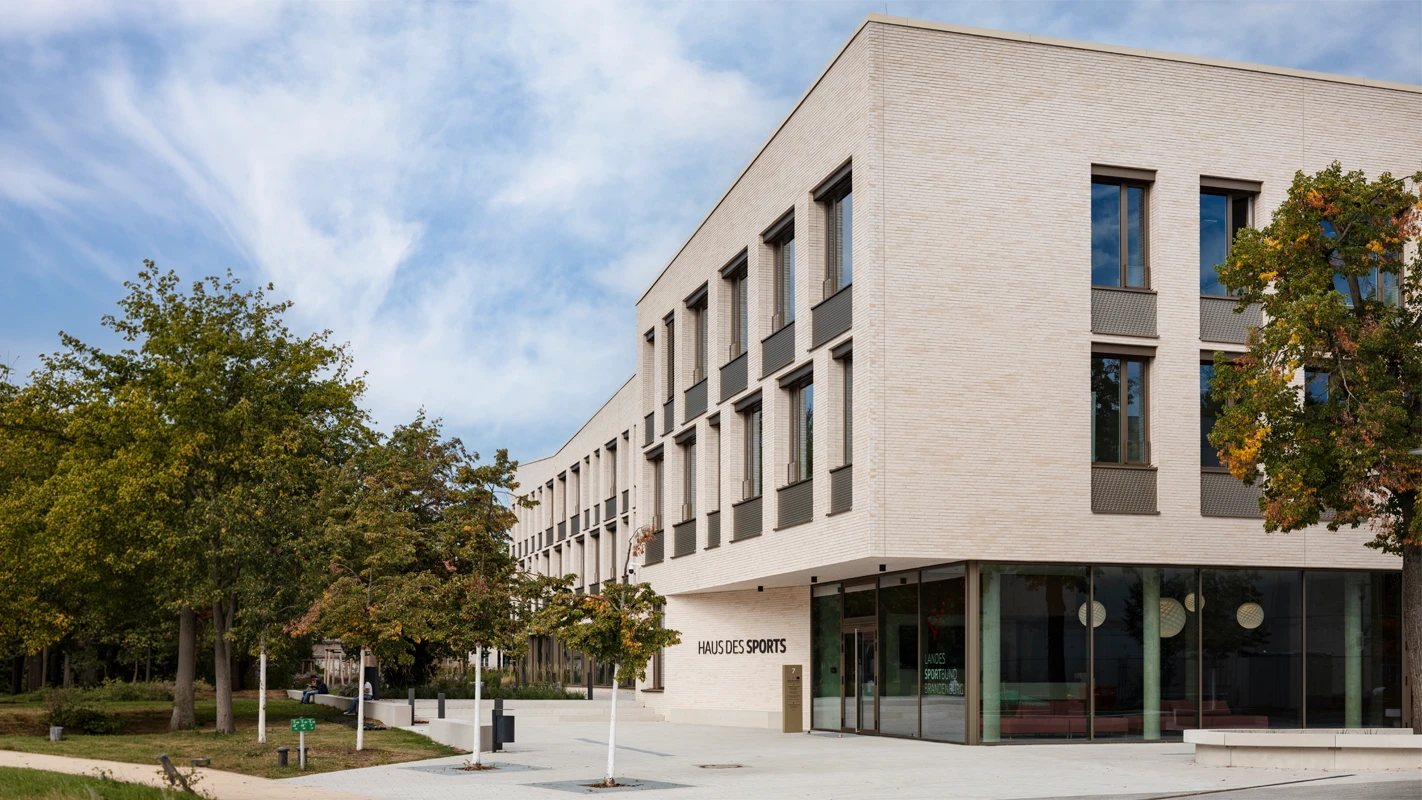Außenansicht des Haus des Sports beim Landessportbund Potsdam mit heller Fassadenbekleidung, großen Glasflächen, 3D-Optik-Platten und modernem Fassadensystem im grünen Umfeld.