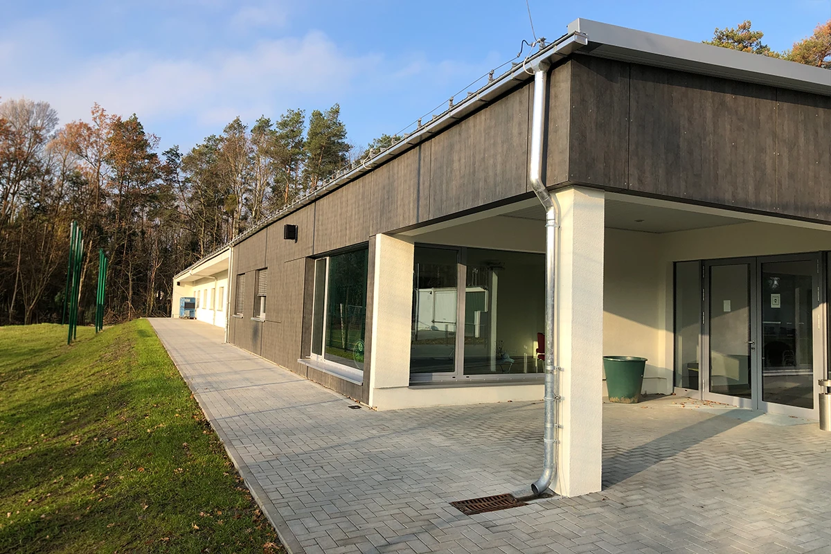 Seitliche Ansicht des Funktionsgebäudes am Sportplatz Michendorf mit dunkler VHF-Fassadenbekleidung, heller Fassade, Glasflächen und gepflastertem Außenbereich nach Fassadensanierung mit WDVS.