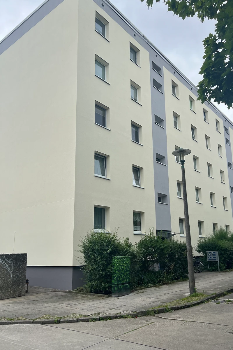Mehrfamilienhaus mit frisch beschichteter heller Fassade, grauem Akzentstreifen und saniertem Sockelbereich nach professioneller Fassadenbeschichtung.