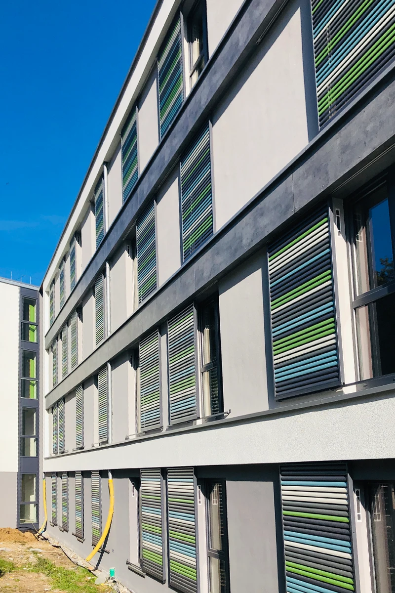 Seitliche Ansicht eines Studentenwohnheims mit moderner Fassade, WDVS, großen Fenstern und farbigen Sonnenschutzelementen nach professioneller Fassadensanierung.